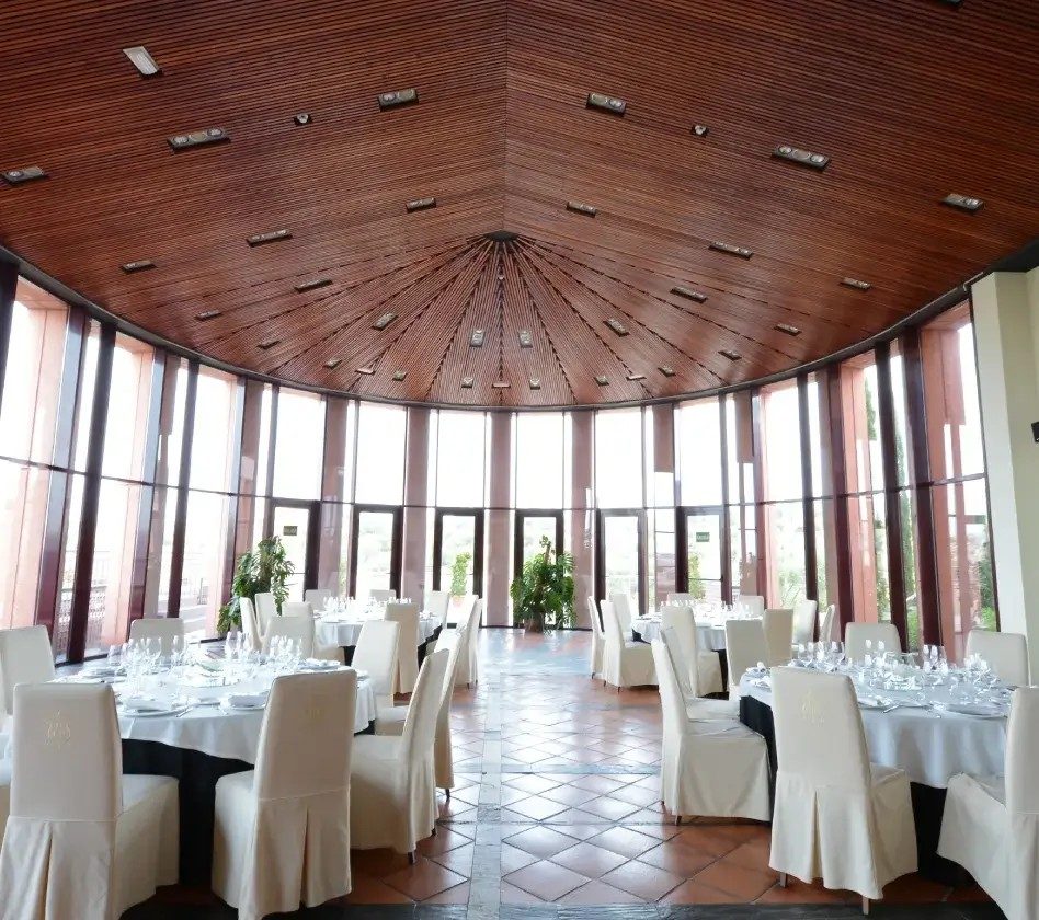 Sala amplia con mesas redondas, sillas blancas, techo de madera y ventanales