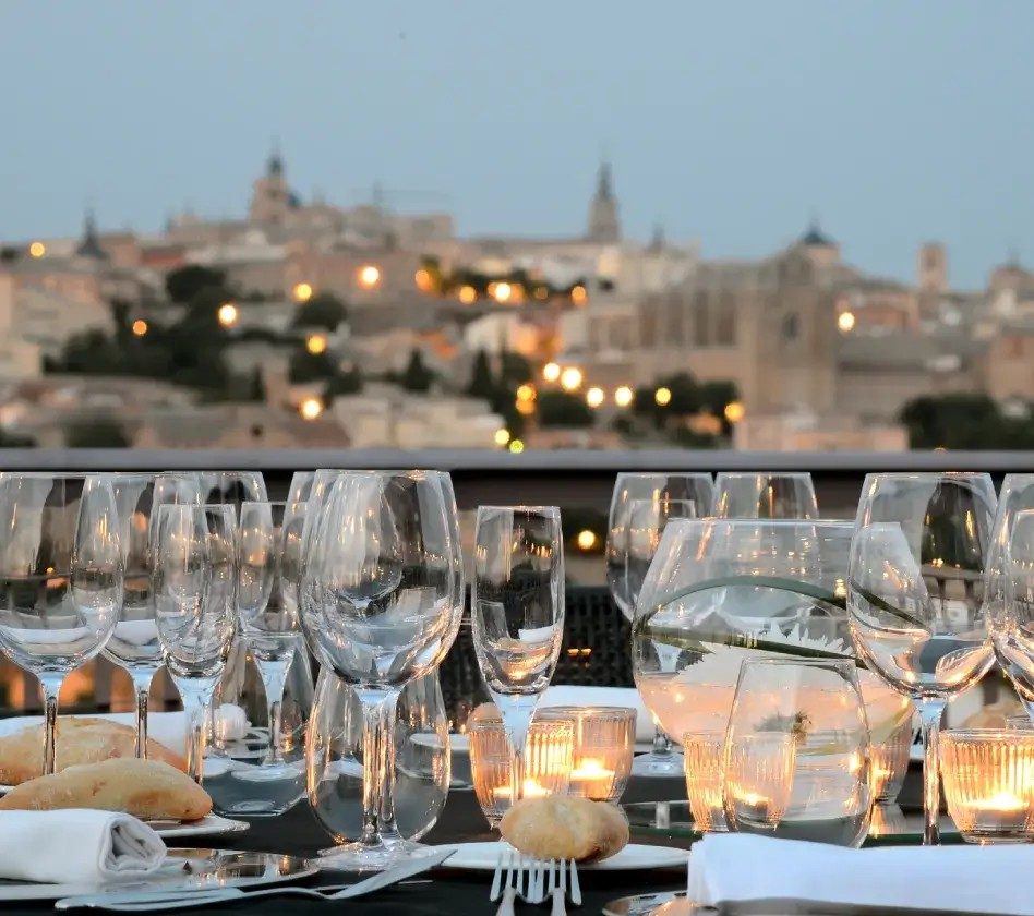 Mesa montada con copas y panecillos, con la ciudad de Toledo iluminada al fondo